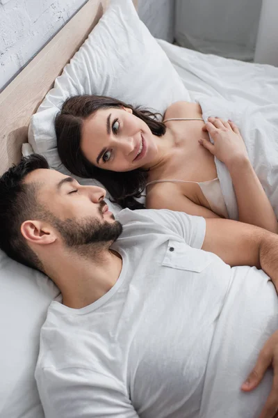 High angle view of smiling young woman looking at sleeping boyfriend in bed with white linen in bedroom — Stock Photo