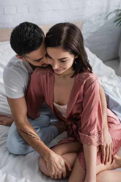 Vista de ángulo alto de la joven sonriente en peignoir sentado en la cama con el novio en el dormitorio — Stock Photo