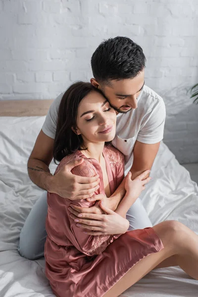 Sonriente joven pareja con los ojos cerrados abrazándose suavemente en el dormitorio - foto de stock