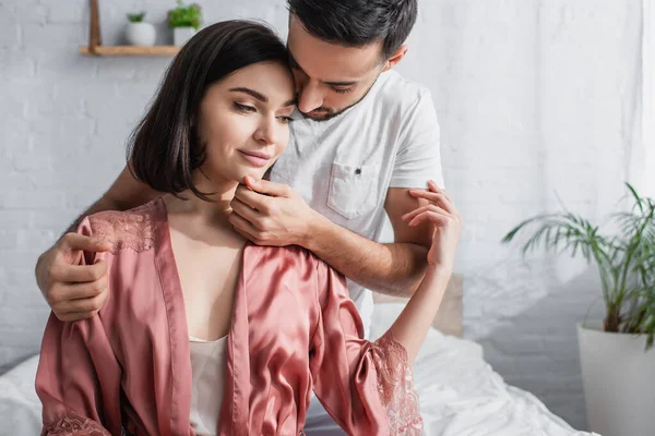 Joven hombre suavemente tocando la cara de la novia en peignoir en el dormitorio - foto de stock