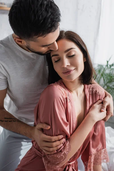 Lächelndes junges Paar mit geschlossenen Augen, das sich im Schlafzimmer sanft umarmt — Stockfoto