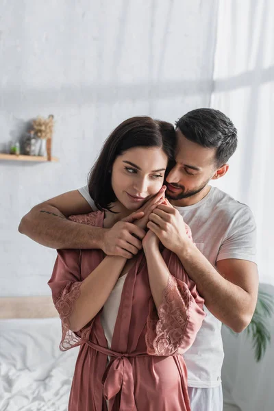 Sonriente joven en peignoir abrazándose y tomándose de la mano con su novio en el dormitorio — Stock Photo
