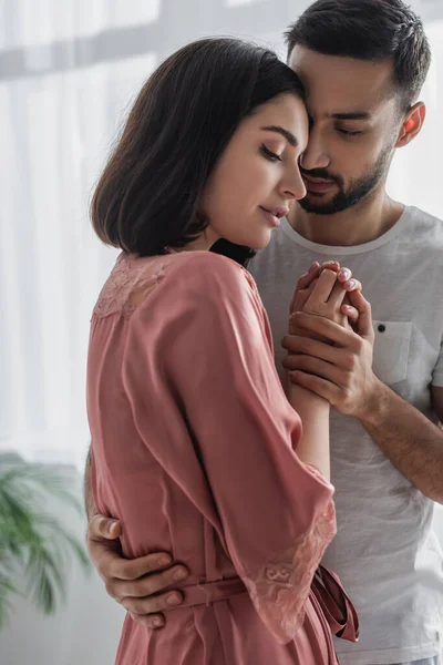 Jovem mulher no peignoir abraçando e de mãos dadas com namorado no quarto — Fotografia de Stock