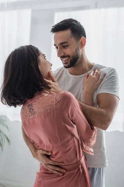 Joven hombre suavemente tocando la cara de la novia en peignoir en el dormitorio - foto de stock