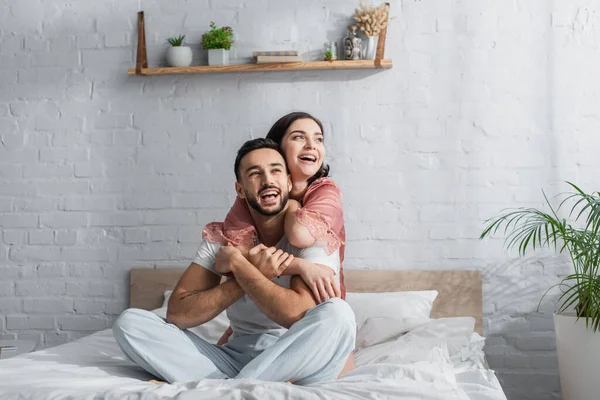 Sonriente joven pareja sentada en la cama con ropa blanca y abrazos en el dormitorio - foto de stock