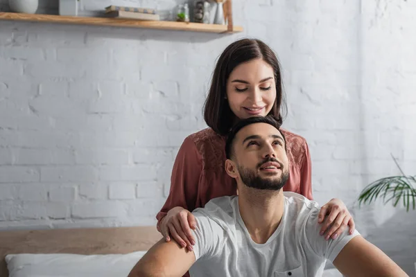 Lächelndes junges Paar sitzt mit weißer Bettwäsche auf dem Bett und umarmt sich im Schlafzimmer — Stockfoto