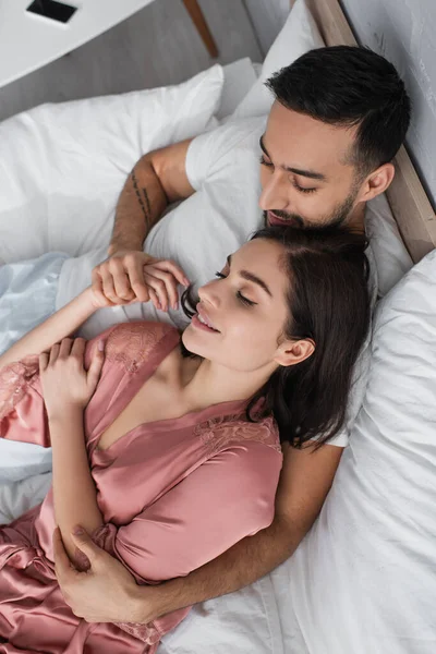 Visão de alto ângulo de casal jovem sorridente deitado na cama com linho branco e de mãos dadas no quarto — Fotografia de Stock