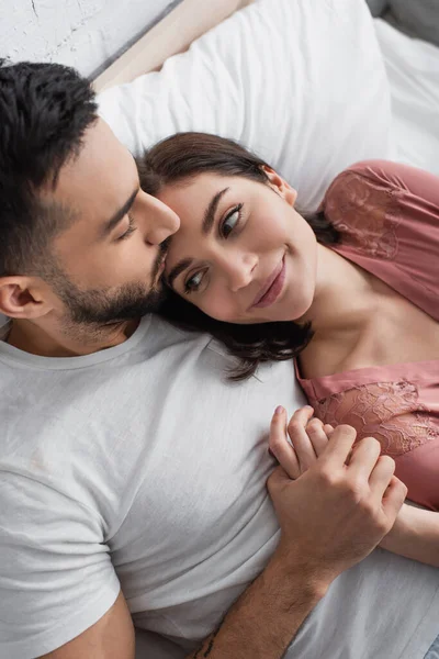 Young man lying on bed with white linen and gently kissing girlfriend in peignoir in bedroom — Stock Photo