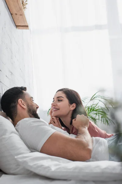 Happy young man lying on bed with white linen and gently touching face of girlfriend in peignoir in bedroom — Stock Photo