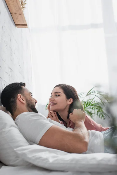 Felice giovane uomo sdraiato sul letto con biancheria bianca e delicatamente toccante viso di fidanzata in peignoir in camera da letto — Foto stock