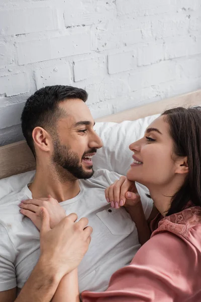 Glücklicher junger Mann liegt mit weißer Bettwäsche auf dem Bett und hält sanft die Hand seiner Freundin im Peignoir im Schlafzimmer — Stockfoto