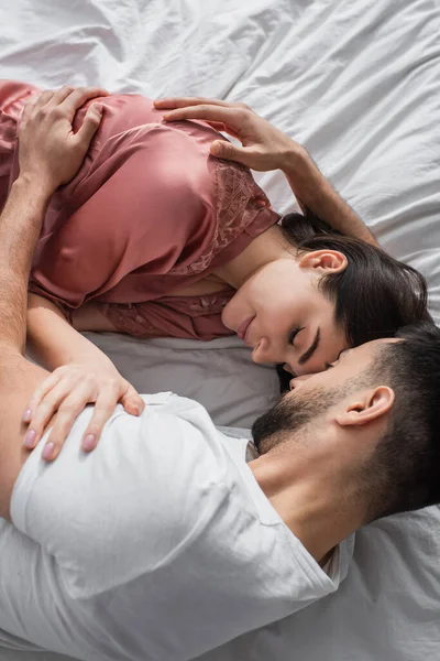 Vista superior do jovem deitado na cama com linho branco e gentilmente abraçando namorada com olhos fechados no quarto — Fotografia de Stock