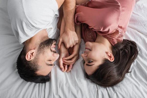 Vista superior do jovem deitado na cama com linho branco e segurando suavemente as mãos da namorada no quarto — Fotografia de Stock