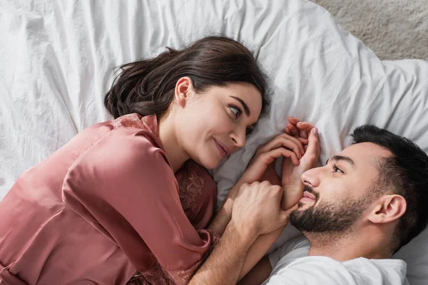Top view of smiling young couple lying on bed with white linen and looking at each other in bedroom — Stock Photo