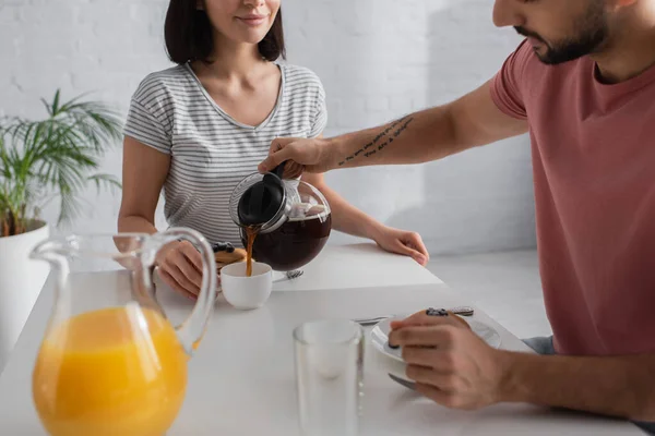 Visão parcial do jovem derramando café de panela a xícara perto da namorada sentada à mesa com café da manhã na cozinha — Fotografia de Stock