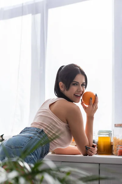 Happy young woman holding orange and looking at camera in kitchen — Stock Photo