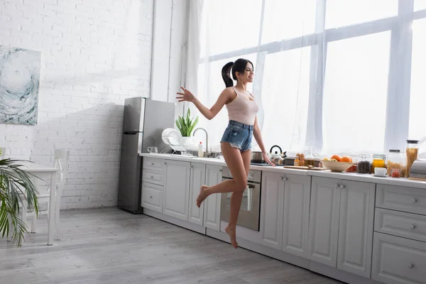 Comprimento total de alegre jovem mulher pulando perto de frutas na cozinha — Fotografia de Stock