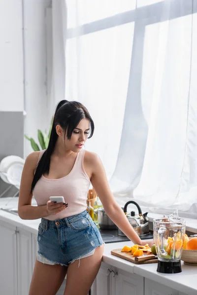 Jovem morena segurando smartphone e olhando para frutas na cozinha — Fotografia de Stock
