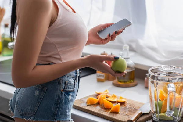 Cropped view of woman holding smartphone and apple in kitchen — Stock Photo