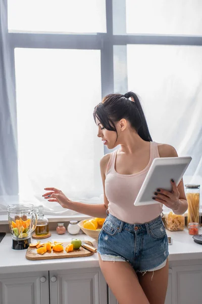 Брюнетка молодая женщина с цифровой планшет и глядя на фрукты на кухне — стоковое фото
