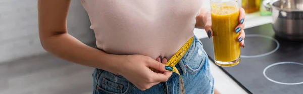 Abgeschnittene Ansicht einer jungen Frau, die ein Glas Smoothie hält, während sie die Taille misst, Banner — Stockfoto