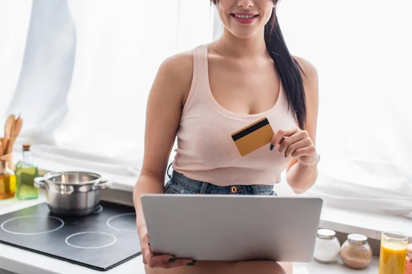 Vue recadrée de la jeune femme tenant la carte de crédit et en utilisant un ordinateur portable dans la cuisine — Photo de stock