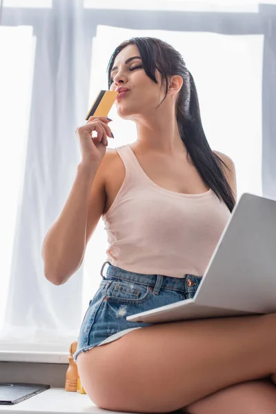 Jovem mulher beijando cartão de crédito e usando laptop na cozinha — Fotografia de Stock