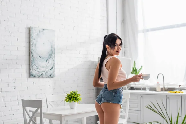 Cheerful young woman holding cup of coffee in kitchen — Stock Photo