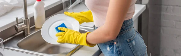 Vista recortada de la mujer joven en guantes de goma lavavajillas en la cocina, pancarta - foto de stock