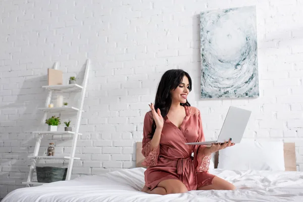 Mujer feliz en bata de seda agitando la mano durante la videollamada en el dormitorio - foto de stock