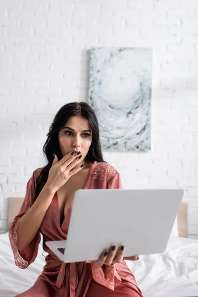 Überraschte junge Frau in Seidenmantel, die Mund bedeckt und auf Laptop schaut — Stockfoto
