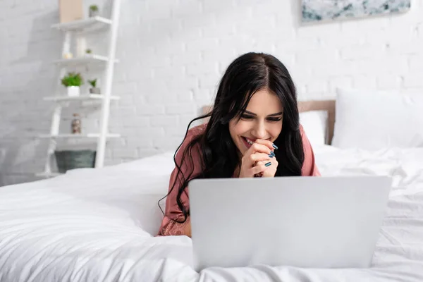 Веселая молодая женщина с сжатыми руками смотрит на ноутбук в спальне — стоковое фото