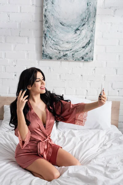 Joven mujer feliz en bata de seda tomando selfie en el teléfono inteligente en el dormitorio - foto de stock