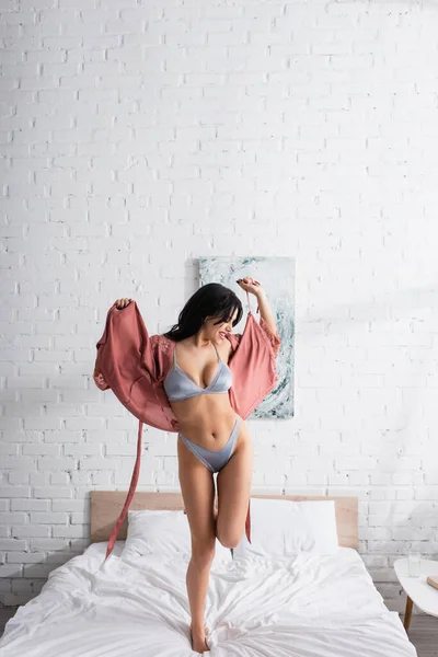 Femme gaie en sous-vêtements sexy et robe de soie dansant sur le lit — Photo de stock