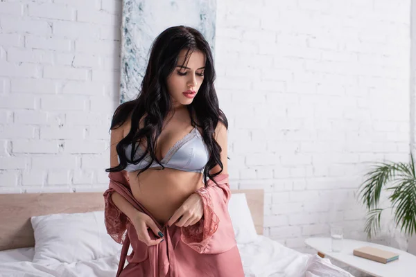 Bruna donna in reggiseno sexy indossa vestaglia di seta mentre seduto sul letto — Foto stock