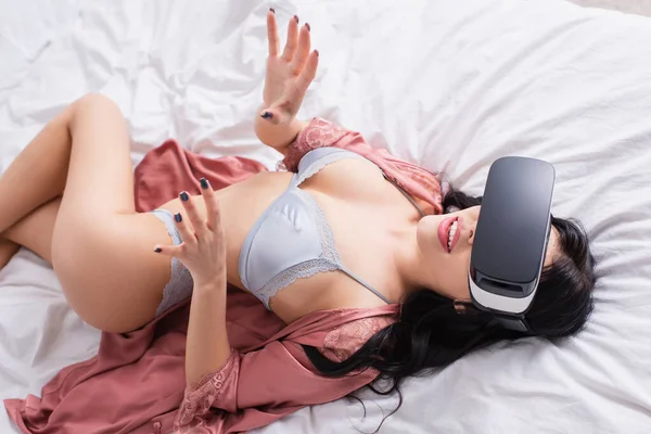 Vista ad alto angolo di stupito giovane donna in auricolare vr e lingerie sexy sdraiato e gesticolare sul letto — Foto stock