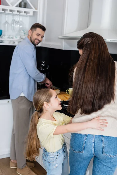 Complacido chica abrazando madre cerca borrosa padre cortar pan en la cocina - foto de stock