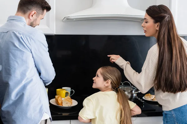 Mujer señalando con el dedo cerca feliz hija y marido preparando el desayuno - foto de stock