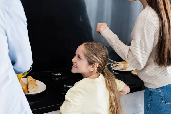 Menina feliz sorrindo perto dos pais preparando café da manhã na cozinha — Fotografia de Stock