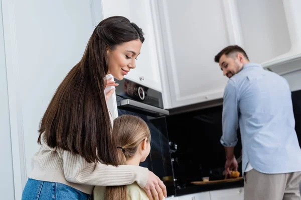 Happy mother hugging child near blurred husband cutting bread in kitchen — Stock Photo
