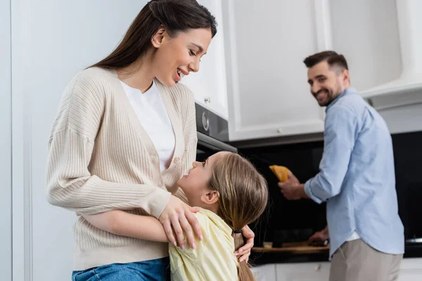 Excitada mujer abrazando hija cerca borrosa marido sosteniendo pan en la cocina - foto de stock