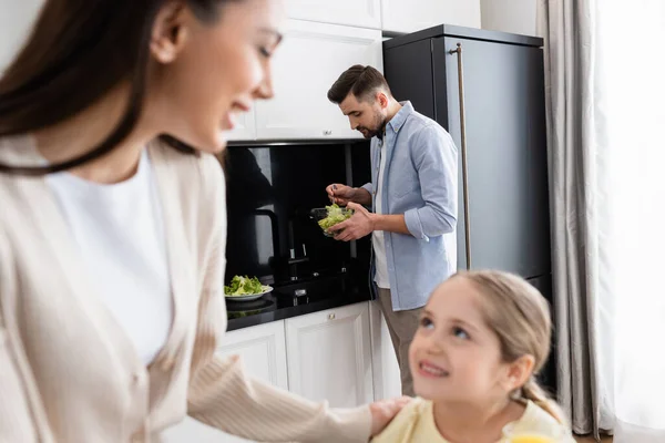 Man preparing vegetable salad near blurred wife and daughter in kitchen — Stock Photo