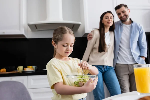 Menina mistura salada de legumes perto de pais felizes no fundo borrado — Fotografia de Stock