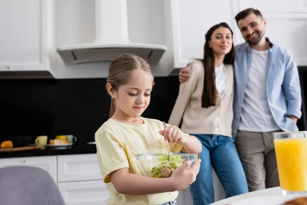 Mädchen mixt Gemüsesalat in der Nähe glücklicher Eltern auf verschwommenem Hintergrund — Stockfoto