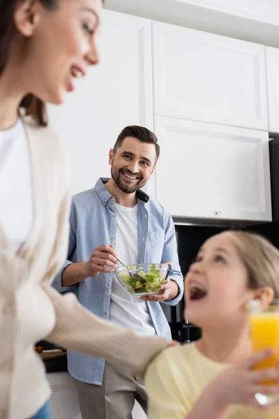 Sonriente hombre mezclando ensalada de verduras cerca borrosa esposa e hija en la cocina - foto de stock