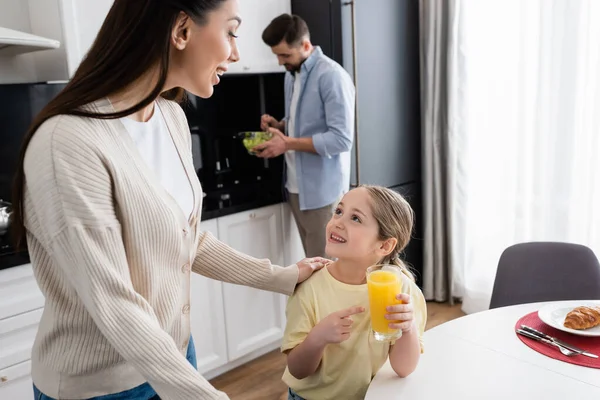 Ragazza allegra che punta al succo d'arancia vicino a mamma e papà mescolando insalata di verdure su sfondo sfocato — Foto stock