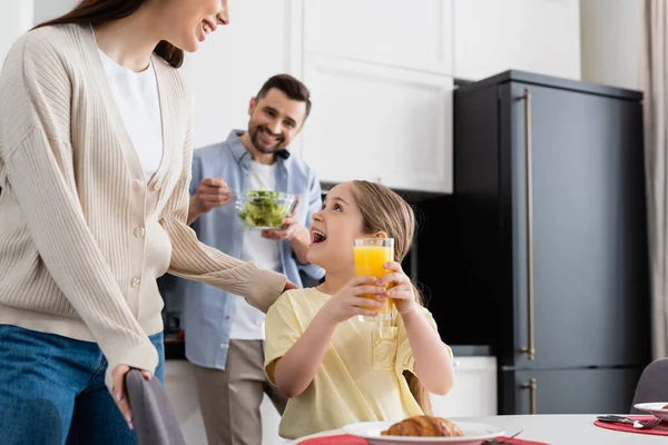 Fille étonnée tenant du jus d'orange près de la mère souriante et le père flou mélange salade dans la cuisine — Photo de stock