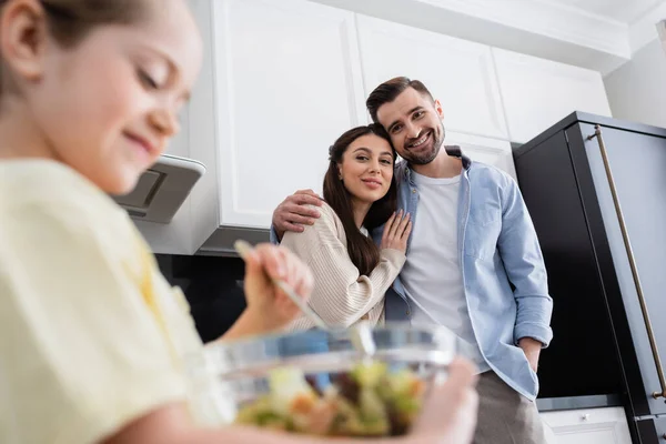 Glückliches Paar umarmt sich in Küche nahe verschwommener Tochter, die Salat in Schüssel mixt — Stockfoto