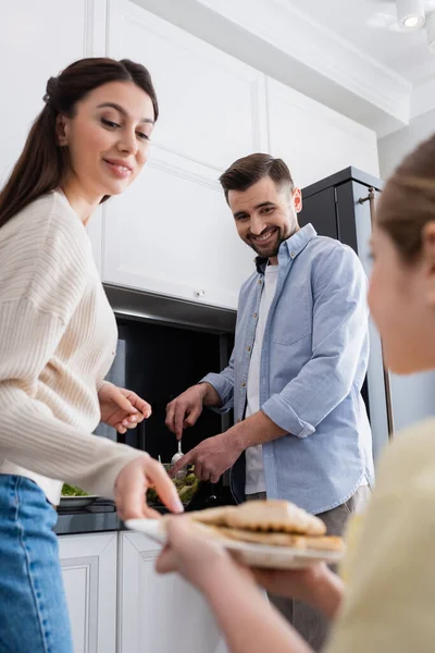 Niño borroso con filete de pollo a la parrilla cerca de mamá y papá sonriente preparando ensalada de verduras - foto de stock