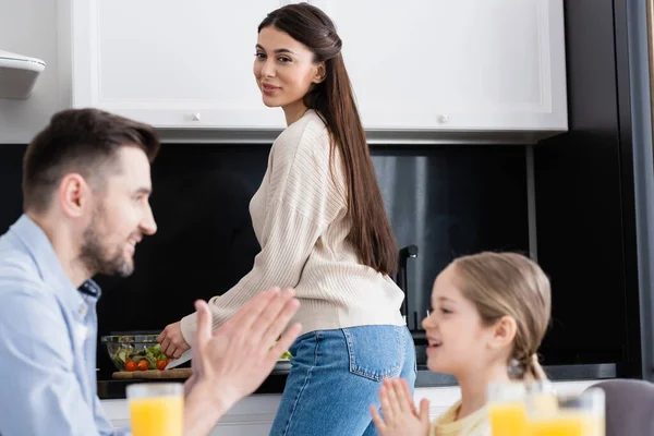 Feliz mujer preparando el desayuno mientras mira borrosa padre e hija jugando pastel de empanada juego - foto de stock
